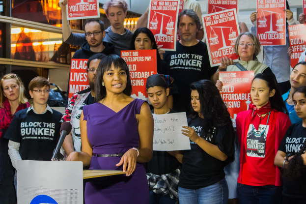 Eine Black-Lives-Matter-Aktivistin steht an einem Podium mit einem Mikrofon und spricht zu einer Menge von Menschen, von denen einige Schilder halten, während im Hintergrund Lichter und andere Gegenstände zu sehen sind.