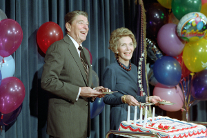 Ein Mann und eine Frau stehen nebeneinander, lächeln und halten jeweils einen Teller in der Hand, vor einem Tisch mit einer Kerzen geschmückten Torte, bunten Ballons und einem Vorhang im Hintergrund, wahrscheinlich feiern sie ihren 60. Geburtstag.
