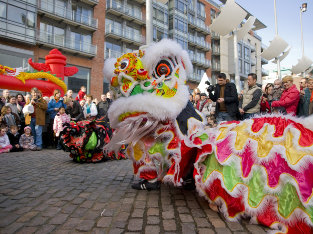 Lebendige Feier des chinesischen Neujahrs in Amsterdam mit einer Löwen-Tanz-Performance vor einer Zuschauermenge, darunter einige mit Kameras, vor einer Kulisse aus Gebäuden, Laternenmasten und einem klaren blauen Himmel.