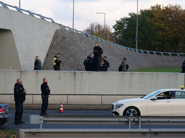 Gruppe von Polizeibeamten steht neben einem Auto auf der Straße, mit Verkehrskegeln, einer Begrenzung, Gras, einer Wand, Laternenmasten, Bäumen und einem bewölkten Himmel im Hintergrund.