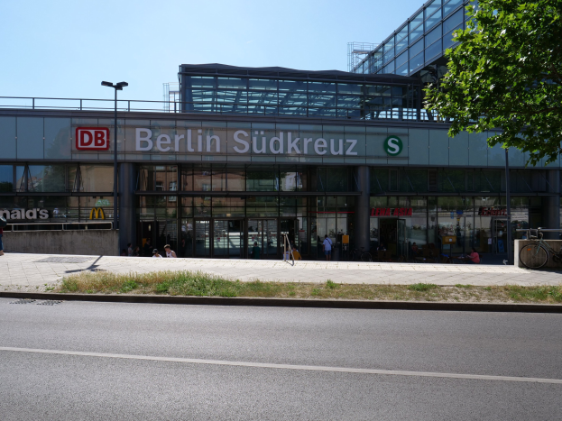 Großes gläsernes Berliner Südkreuz-Bahnhof mit Text an seiner Fassade, umgeben von Straßenlaternen, Lichtern, Fahrzeugen, Fußgängern, Fahrrädern, Bäumen und einem klaren blauen Himmel.