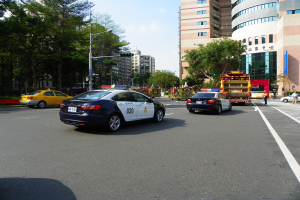 Ein Polizeiwagen fährt auf einer Straße mit hohen Gebäuden, Bäumen, Laternen, Ampeln und Menschen unter einem bewölkten Himmel.