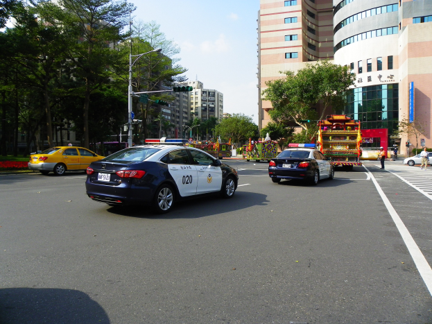 Ein Polizeiwagen fährt auf einer Straße mit hohen Gebäuden, Bäumen, Laternen, Ampeln und Menschen unter einem bewölkten Himmel.