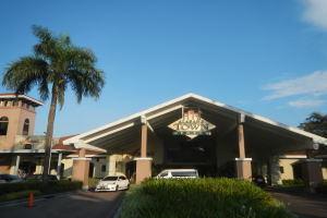 Der Eingang zu einem Resort mit Palmen vorne, umgeben von Gebäuden, Kraftfahrzeugen auf der Straße, Verkehrskegeln, Büschen, Bäumen und einem bewölkten Himmel im Hintergrund.