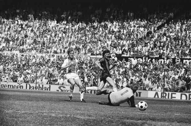 Schwarzes Foto von Männern, die auf einem Feld Fussball spielen, mit Zuschauern im Hintergrund und einer "Bundesliga 1978-79 Ajax 1-1 Hannover 96" Fahne.