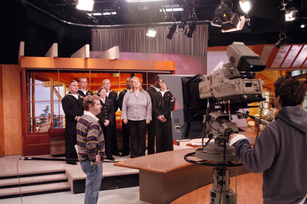 Gruppe von Menschen in einem Fernsehstudio mit einer Person, die eine Kamera hält, auf der rechten Seite und Deckenlampen im Hintergrund.
