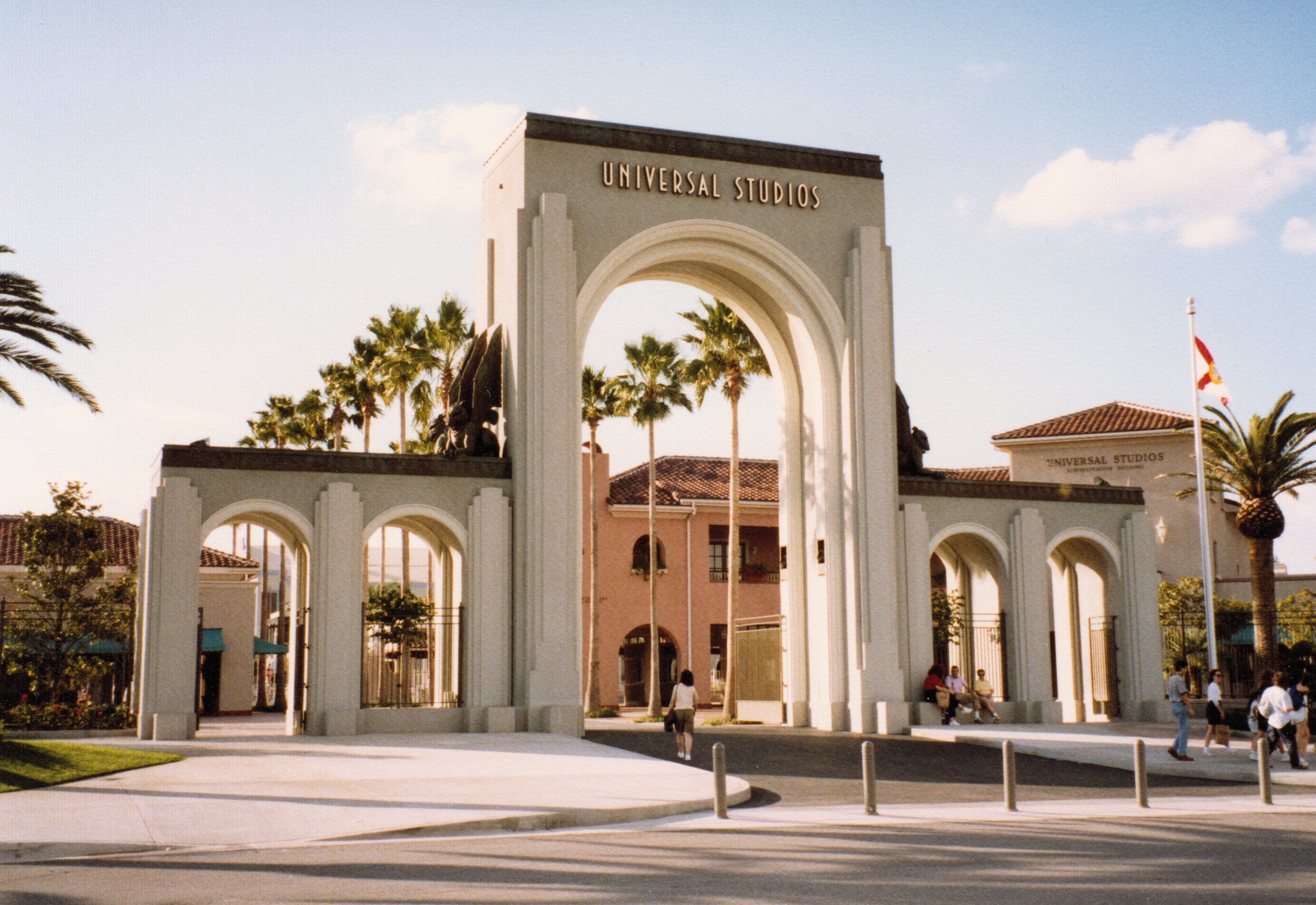 Eingang von Universal Studios Hollywood mit einem Bogen, einer Statue, Menschen, Bäumen, Gebäuden, einem Fahnenmast mit einer Fahne und einem bewölkten Himmel.