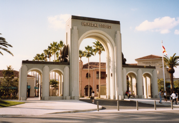 Eingang von Universal Studios Hollywood mit einem Bogen, einer Statue, Menschen, Bäumen, Gebäuden, einem Fahnenmast mit einer Fahne und einem bewölkten Himmel.