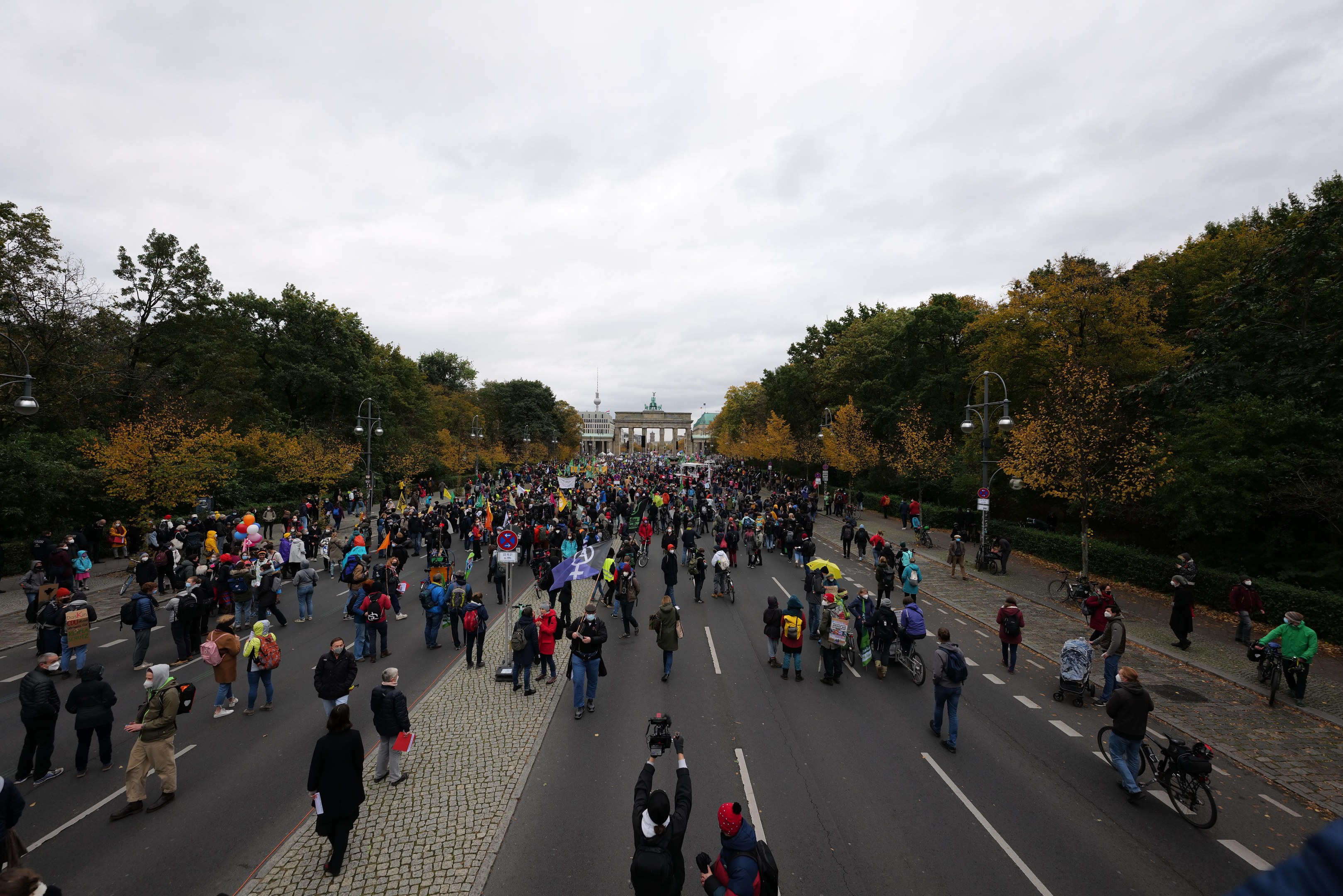 Eine große Gruppe von Menschen marschiert eine von Bäumen gesäumte Straße in Berlin entlang, mit Kameras und Schildern in der Hand, vor einem Gebäude und einem klaren Himmel.