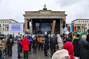 Eine Menschenmenge steht vor einem Gebäude mit einer Bühne, Lautsprechern und einem Bildschirm, mit Flaggen und Transparenten auf der rechten Seite, was auf eine Protestaktion in Berlin hindeutet.