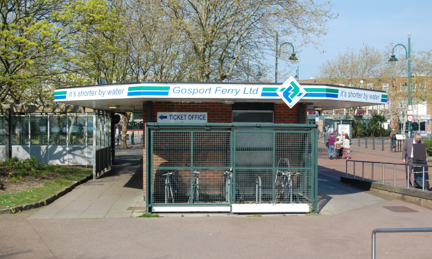 Bus-Haltestelle mit 'Gosport Ferry Ltd'-Schild, Metallzaun, Menschen, Fahrrädern, Bäumen, Straßenlaternen, Schildern, Gebäude und bewölktem Himmel.