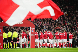 Eine Gruppe von Menschen auf einem Fußballfeld mit einer roten und weißen Flagge im Vordergrund, einem Bogen mit der Aufschrift "Bayern München vs Bayern München Wetten & Vorschau" im Hintergrund und einer großen Menge im Stadion.