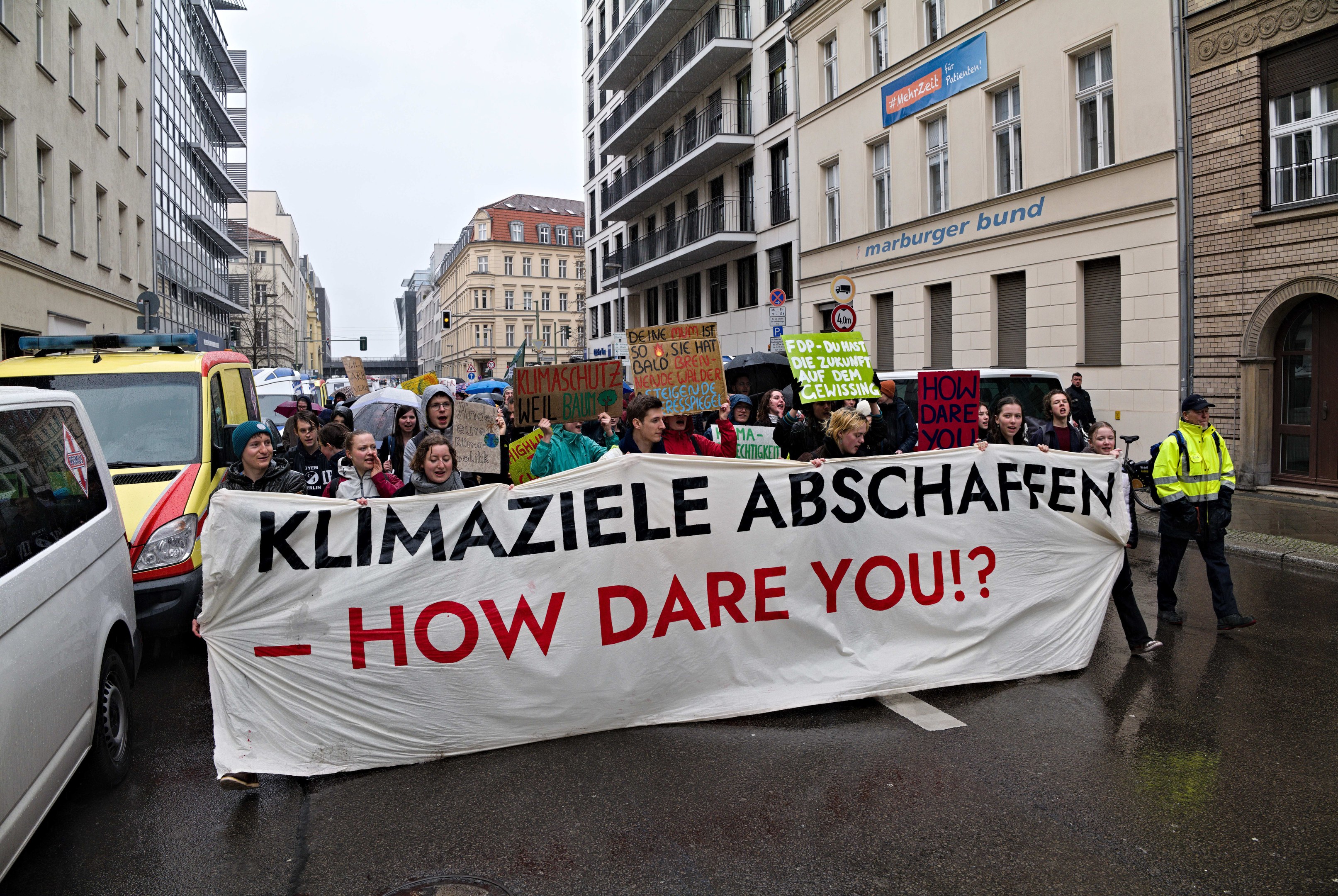 Gruppe von Menschen mit einem Banner "Klimaziele Abschaffen - Wie Wägen Sie?" auf der Straße unterwegs mit Fahrzeugen, Gebäuden, Schildern und einem klaren blauen Himmel.
