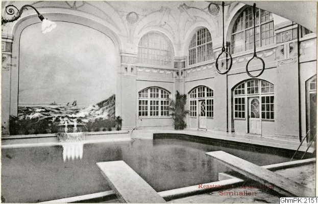 Altes Schwarz-Weiß-Foto eines großen Hallenbades in einem Gebäude mit Glasfenstern und -türen, umgeben von Pflanzen und einer Deckenlampe, beschriftet mit "Rensburg, Deutschland - Schwimmbad".