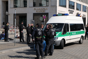 Eine Gruppe von Polizeibeamten steht vor einem Starbucks-Café, rechts ein Van und links einige Personen; im Hintergrund sind ein Gebäude, ein Schild, ein Laternenmast und ein Zaun zu sehen.