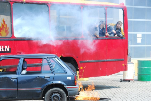 Roter Doppeldeckerbus mit Rauchentwicklung und drei sichtbaren Passagieren, daneben geparktes Auto und ein Gebäude mit Glasfenstern und einem Fass im Hintergrund.