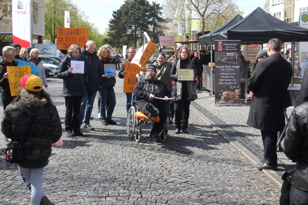 Eine Gruppe von Menschen, die eine Kopfsteinpflasterstraße entlanggehen, einige halten Schilder, mit einer Person im Rollstuhl im Vordergrund und Zelten, Fahrzeugen, Bäumen, Gebäuden, Flaggen und einem klaren blauen Himmel im Hintergrund.