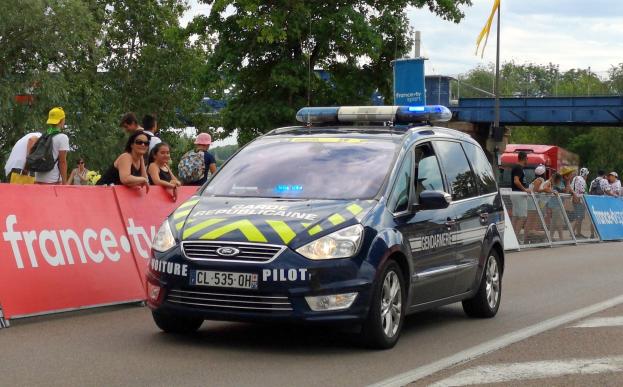 Polizeiauto fährt auf einer Straße neben einer Menschenmenge, mit einem Banner auf der linken Seite, einem Geländer mit Bannern dahinter und Bäumen, einer Brücke, einer Fahne und Wolken im Hintergrund.