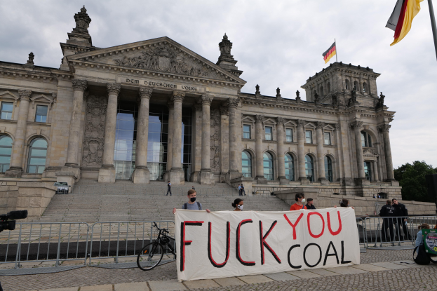 Eine Gruppe von Menschen hält ein "Fuck You Coal"-Schild vor dem Reichstag in Berlin, Deutschland, mit Bäumen, einer Fahnenstange und einem bewölkten Himmel im Hintergrund.