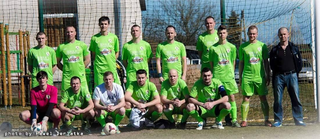 Eine Gruppe von Männern in grünen T-Shirts steht auf einem Fußballfeld mit einem Ball und einem Tor, einige sind in der Hocke, mit Bäumen, Gebäuden und Fahrzeugen im Hintergrund.