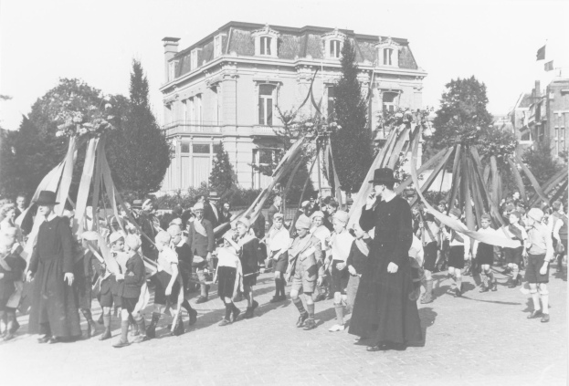 Schwarzes und weißes Bild einer Gruppe von Menschen, die eine Straße entlanggehen, einige halten Fahnen, mit Hüten, einem Maibaum, Bäumen, Gebäuden und Himmel.