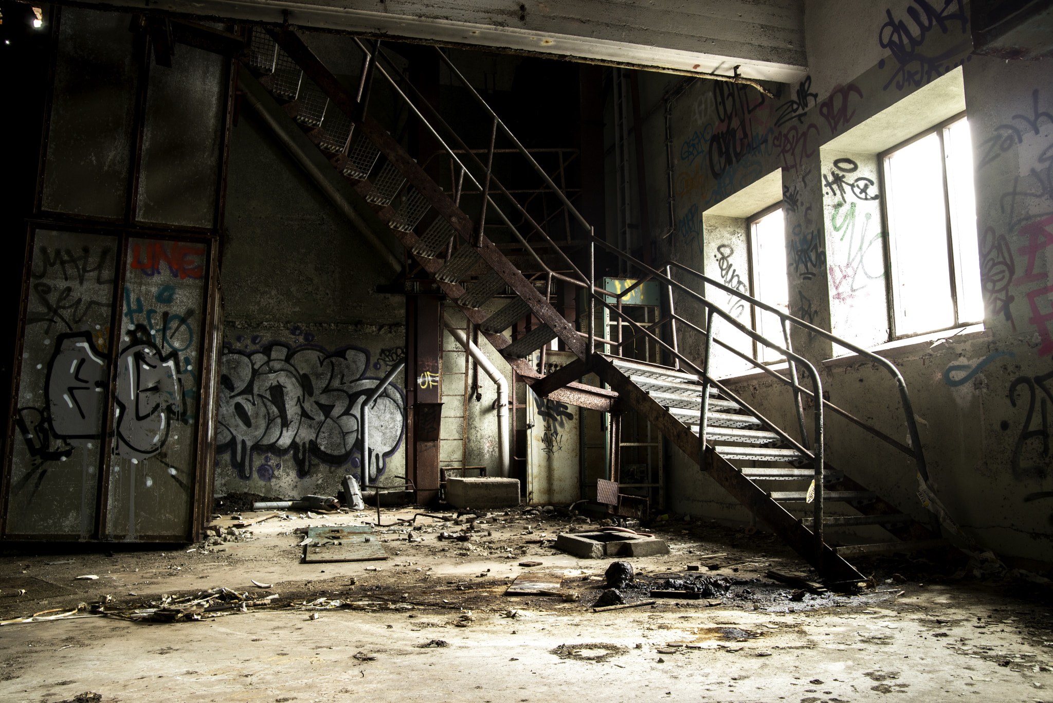 Verlassenes Gebäude mit graffiti-bedeckten Wänden, Treppe mit Geländern, die zum zweiten Stock führen, Fenster auf der rechten Seite, eine Tür auf der linken Seite und verstreute Gegenstände auf dem Boden.