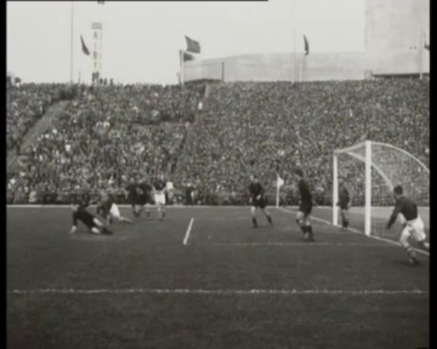 Schwarzes und weißes Foto des WM-Finales 1958 zwischen Manchester United und Liverpool, das Spieler auf dem Feld, ein Tor auf der rechten Seite, Zuschauer in den Rängen und Fahnen im Hintergrund zeigt.