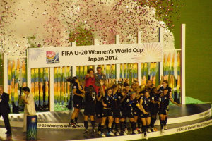 Gruppe von Frauenfußballspielerinnen in schwarzen Uniformen, die auf einer Bühne umgeben von einer Menge feiern, mit einem Hintergrundboard mit Text und Bildern.