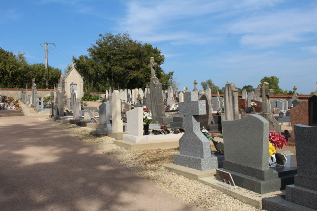 Friedhof mit zahlreichen Grabsteinen und Blumen im Vordergrund, eine Straße im Hintergrund, Bäume, ein Strommast mit Drähten und ein bewölkter Himmel.