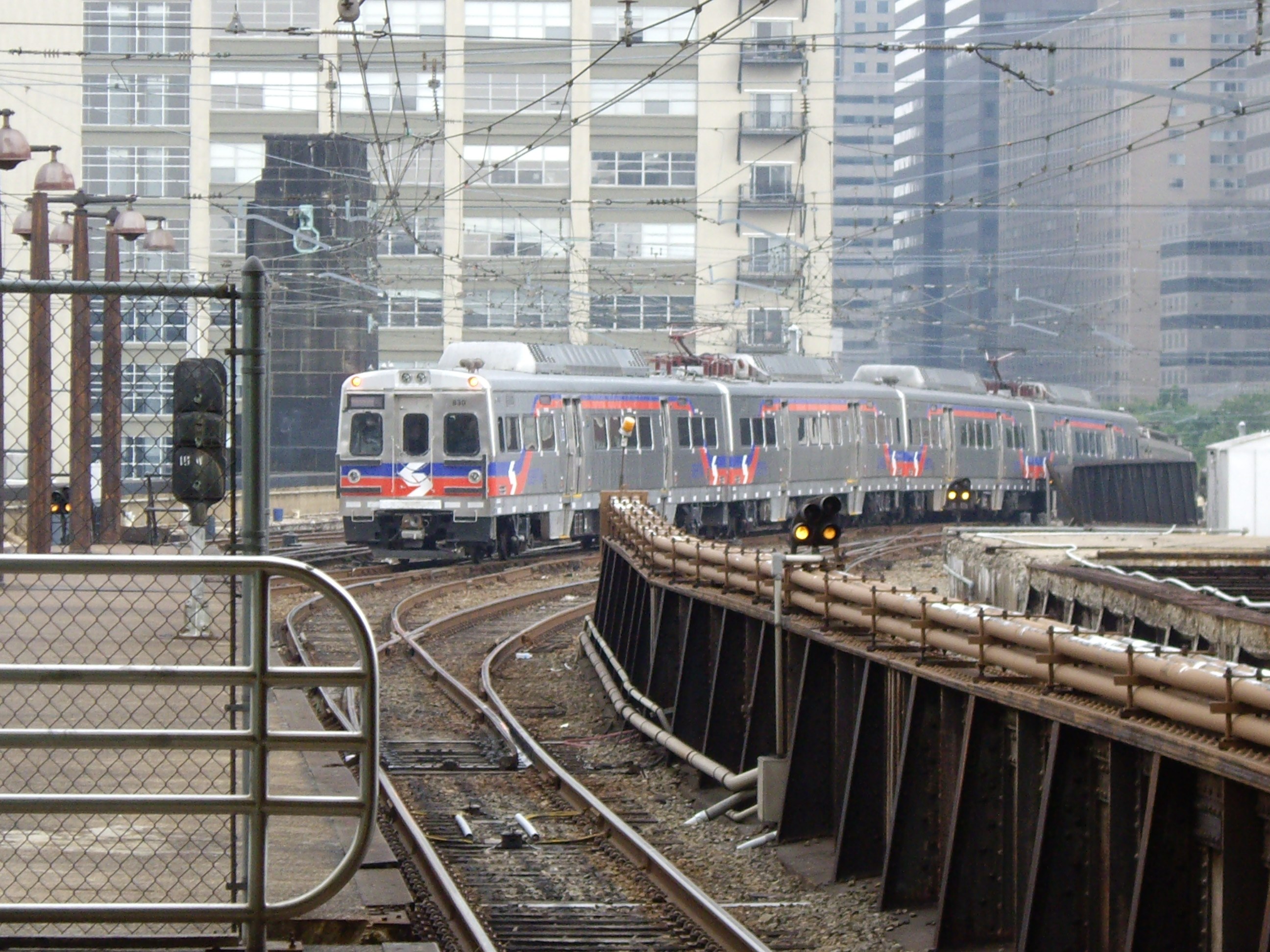 Nahverkehrszug fährt auf Gleisen neben Hochhäusern mit Bahninfrastruktur und städtischen Elementen im Hintergrund.