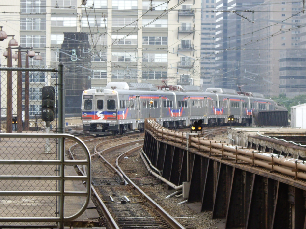 Nahverkehrszug fährt auf Gleisen neben Hochhäusern mit Bahninfrastruktur und städtischen Elementen im Hintergrund.