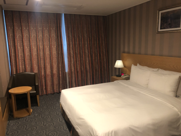 Hotelzimmer mit großem Bett, Stuhl, Tisch, Telefon, Lampe, Foto auf dem Nachttisch, Fenster mit Vorhängen und Deckenleuchten.