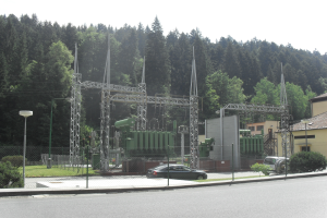 Ein elektrisches Umspannwerk am Straßenrand, eingezäunt und umgeben von Masten, Lampen, Pflanzen, Gras und Bäumen, mit einem vorbeifahrenden Auto und Gebäuden im Hintergrund unter einem klaren blauen Himmel.