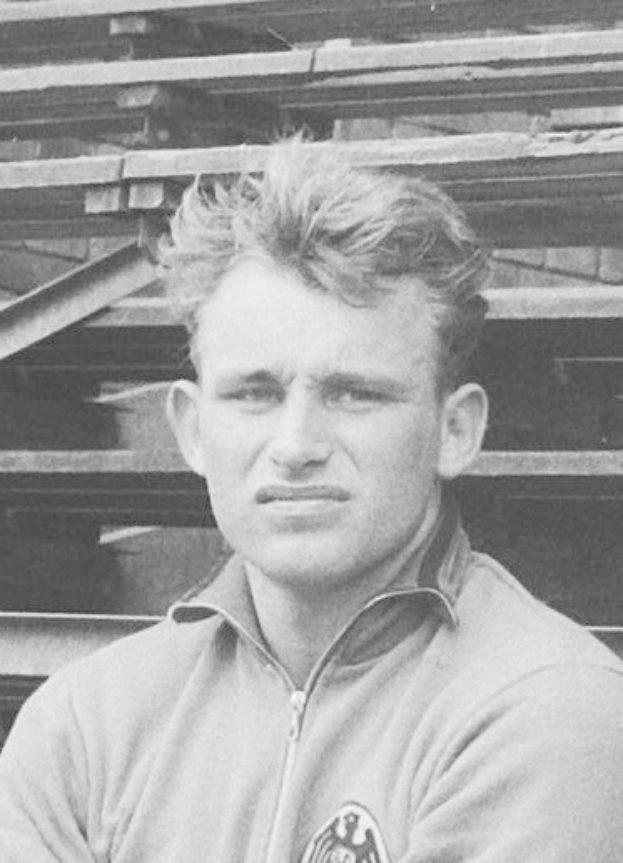 Schwarzes und weißes Foto eines Mannes in einer Jacke vor gestapelten Holzplanken, der als legendärer deutscher Fußballspieler gilt.