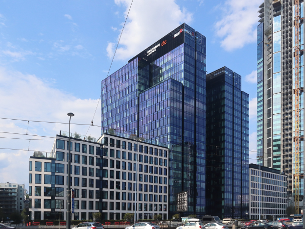 Außenansicht des Deutschen Bank-Hauptsitzes in Frankfurt, Deutschland, mit einem mehrstöckigen Gebäude mit zahlreichen Fenstern, umgeben von städtischer Infrastruktur wie Straßenlaternen, Elektromasten, Elektrokabeln, Fahrzeugen auf der Straße, Büschen, Bäumen und einem wolkenverhangenen Himmel.