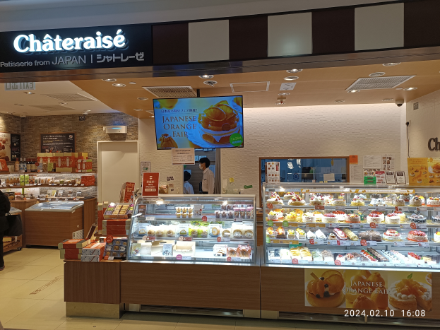 Eine Chateraise-Bäckerei in Japan mit einer Vitrine voller Kuchen und Torten, Menschen stehen und sitzen in der Nähe und Schilder mit Schrift an der Wand im Hintergrund.