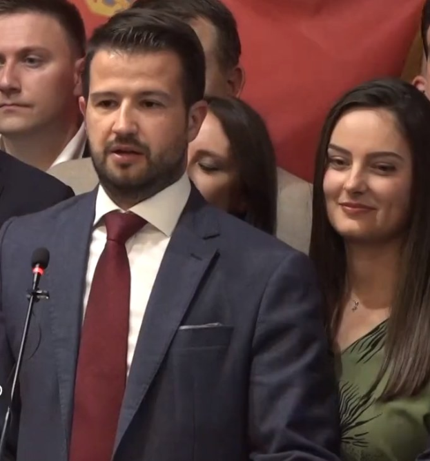 Ein Mann in Anzug und Krawatte spricht vor einem Publikum an einem Rednerpult, mit einem Banner im Hintergrund.