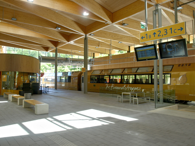 Ein belebter Bahnhof mit einem stehenden Zug auf den Schienen, umgeben von Bänken, Tischen, Mäständen, Schildern, Lichtern und Bäumen, mit wartenden Menschen.