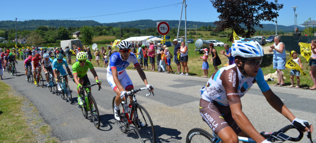 Eine Gruppe von Radfahrern fährt eine Straße entlang, während Zuschauer am Straßenrand stehen, einige tragen Helme und Schutzbrillen, andere halten Fahnen, vor einem Hintergrund aus Bäumen, Schildern, Fahrzeugen, Zelten, Gebäuden, Hügeln und einem klaren blauen Himmel.
