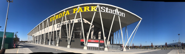 Großes Stadiongebäude mit Text, umgeben von Pfählen, Lichtern, Bäumen und einem Zaun.