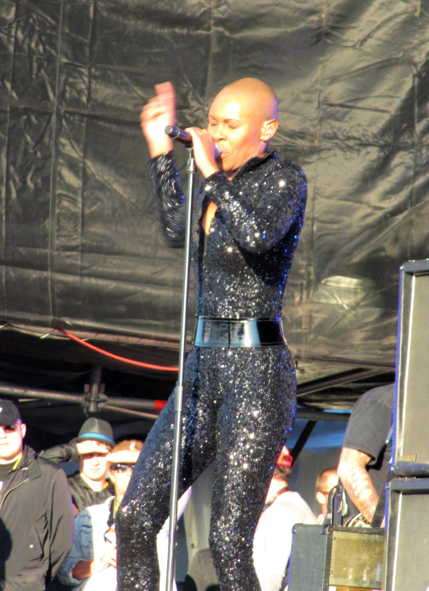 Eine Frau in einem schwarzen Anzug singt in ein Mikrofon auf der Bühne, während eine Gruppe von Menschen mit Mützen und Schutzbrillen hinter ihr Instrumente spielt, mit Lautsprechern und einem schwarzen Tuch im Hintergrund.