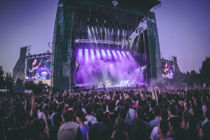 Eine große Menschenmenge auf einem Musikfestival mit einer Bühne, auf der Musiker spielen, beleuchtet von Scheinwerfern und umgeben von Bildschirmen, unter einem klaren blauen Himmel mit Bäumen und einem Gebäude im Hintergrund.