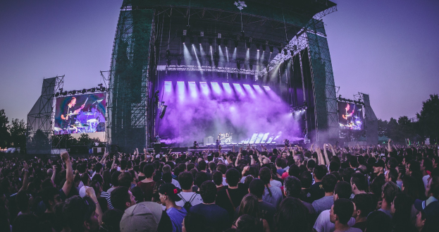 Eine große Menschenmenge auf einem Musikfestival mit einer Bühne, auf der Musiker spielen, beleuchtet von Scheinwerfern und umgeben von Bildschirmen, unter einem klaren blauen Himmel mit Bäumen und einem Gebäude im Hintergrund.