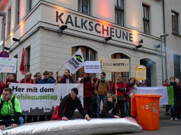 Eine Gruppe von Menschen protestiert vor einem Gebäude in Deutschland, sie halten Schilder und Plakate in den Händen, zwei Personen sitzen im Vordergrund und ein Müllcontainer auf der rechten Seite.