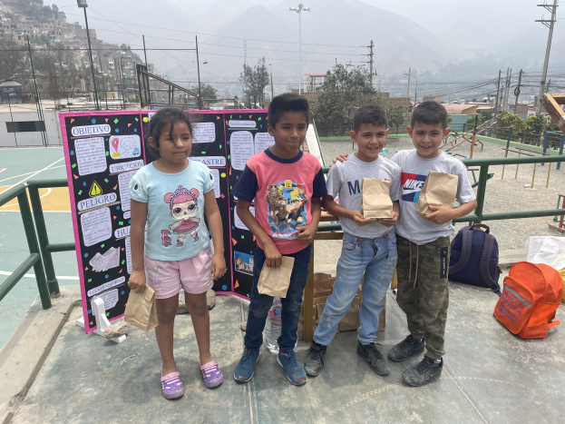 Gruppe von Kindern auf einem Basketballfeld, die Papiertüten halten, mit Gegenständen rechts und einer Tafel mit Text links, vor einer Kulisse aus Zäunen, Pfosten und Gebäuden