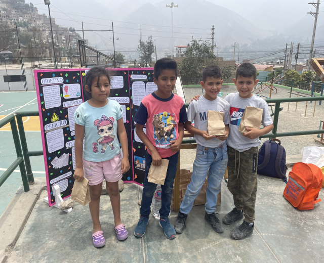 Gruppe von Kindern auf einem Basketballfeld, die Papiertüten halten, mit Gegenständen rechts und einer Tafel mit Text links, vor einer Kulisse aus Zäunen, Pfosten und Gebäuden