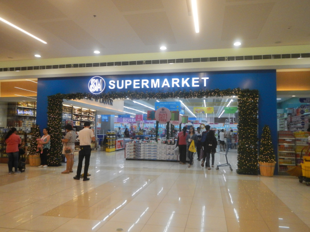 Gruppe von Menschen vor einem Supermarkt mit Einkaufstägern, Taschen und einem Einkaufswagen, Topfpflanze, Regalen, Schildern und Deckenbeleuchtung.