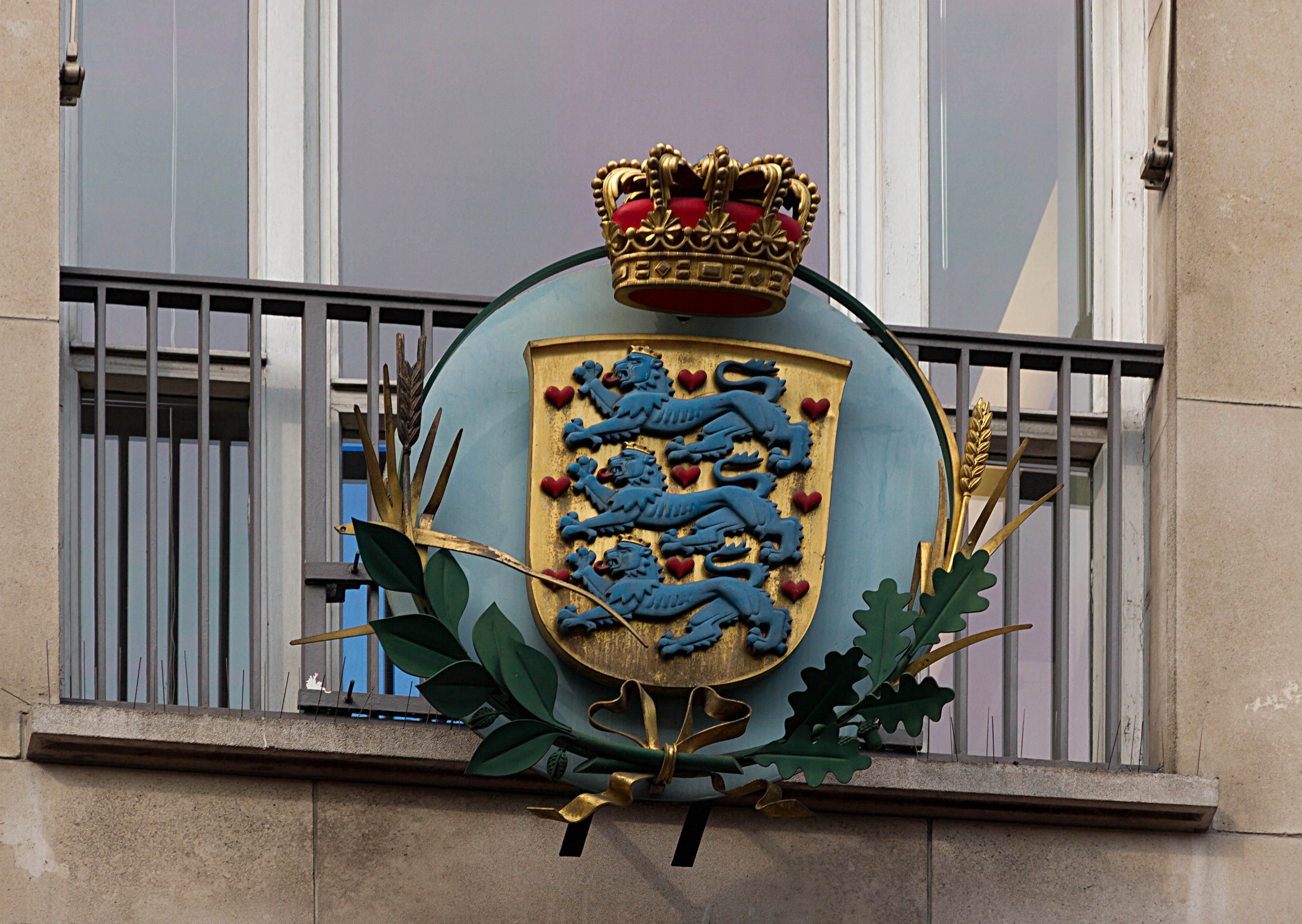 Wappen der britischen Königsfamilie an einer Gebäudewand, mit einer Krone und einem Blatt-Logo, Fenstern und Gittern im Hintergrund.