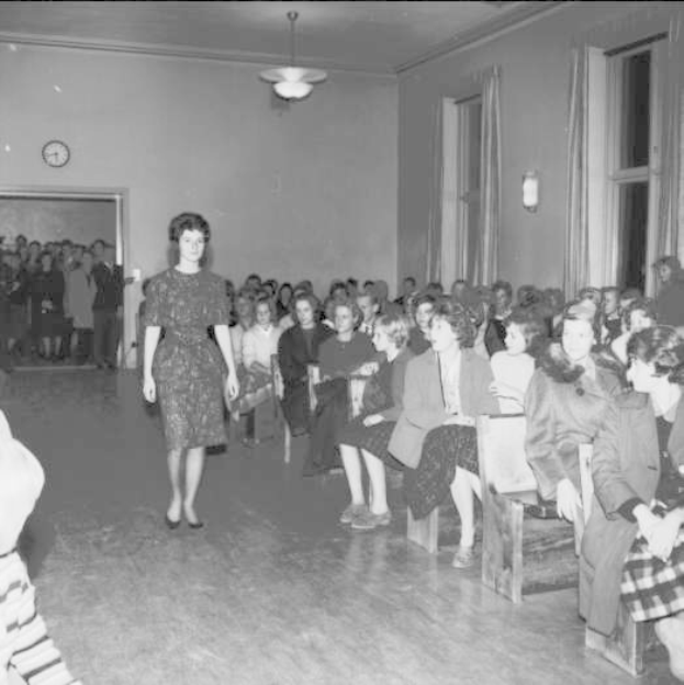 Eine Frau in einem schwarzen und weißen Kleid läuft über den Laufsteg während einer Modenschau für Frauenrechte, mit einer Menge, die auf Stühlen sitzt und einer Uhr und Lampen an der umliegenden Wand.
