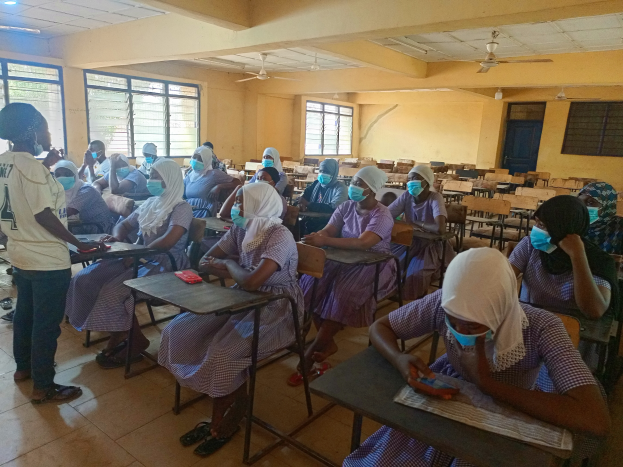 Ein Klassenzimmer in Nigeria mit Schülern, die Gesichtsmasken tragen und auf Bänken und dem Boden sitzen, mit Büchern und Gegenständen auf Tischen, Fenstern, einer Tür, einer Wand und einer Decke mit Lichtern und Ventilatoren im Hintergrund.